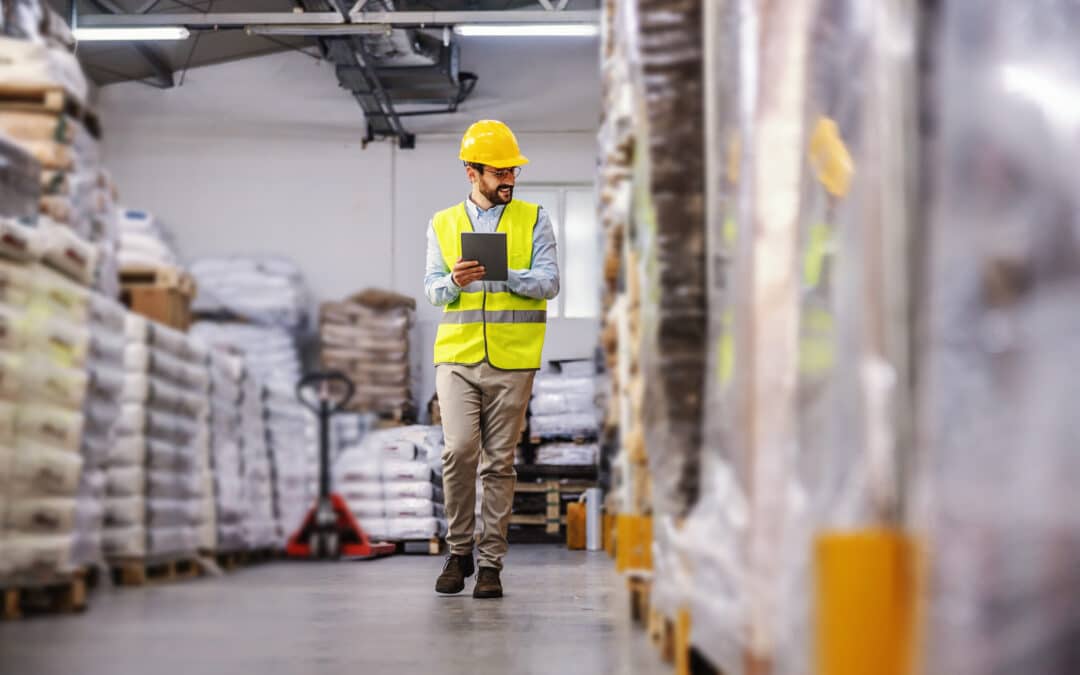 Warehouse worker in safety vest and hard hat reviewing inventory on a tablet while walking through a storage aisle
